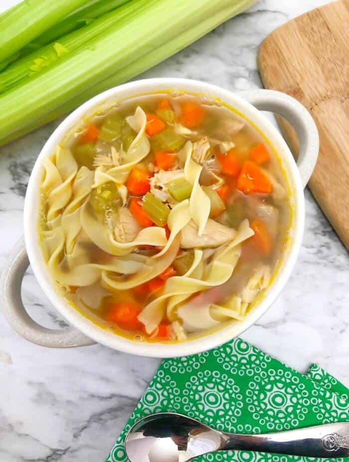 A bowl of chicken noodle soup from overhead with a napkin and celery nearby