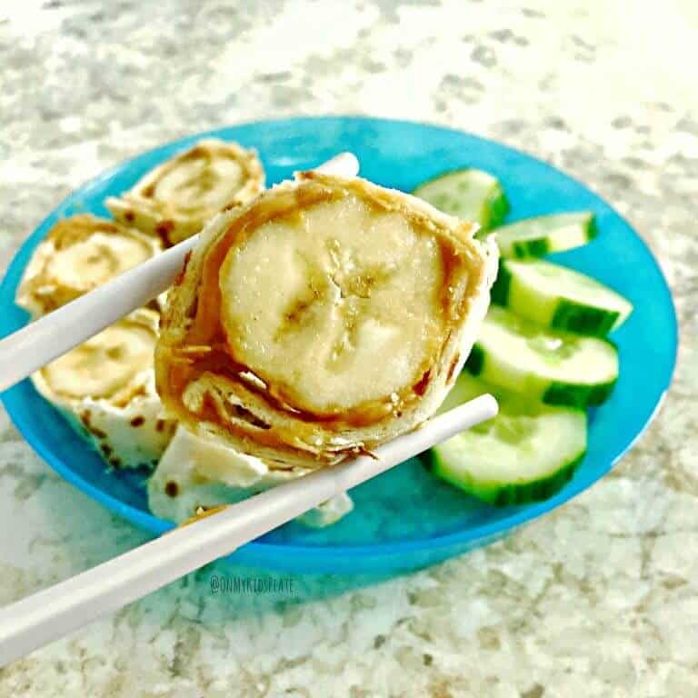 A slice of banana wrapped in peanut butter and tortilla being held by chopsticks over a plate