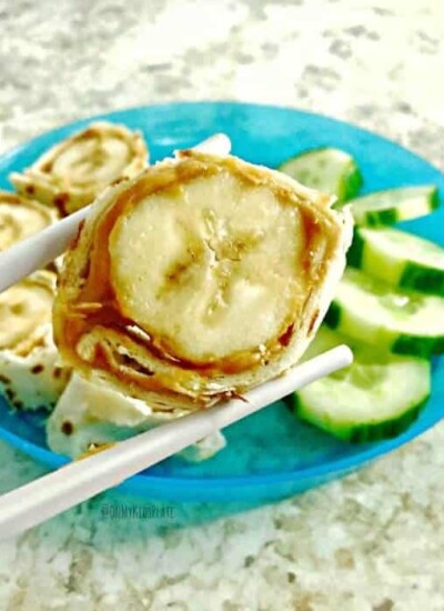 A slice of banana wrapped in peanut butter and tortilla being held by chopsticks over a plate