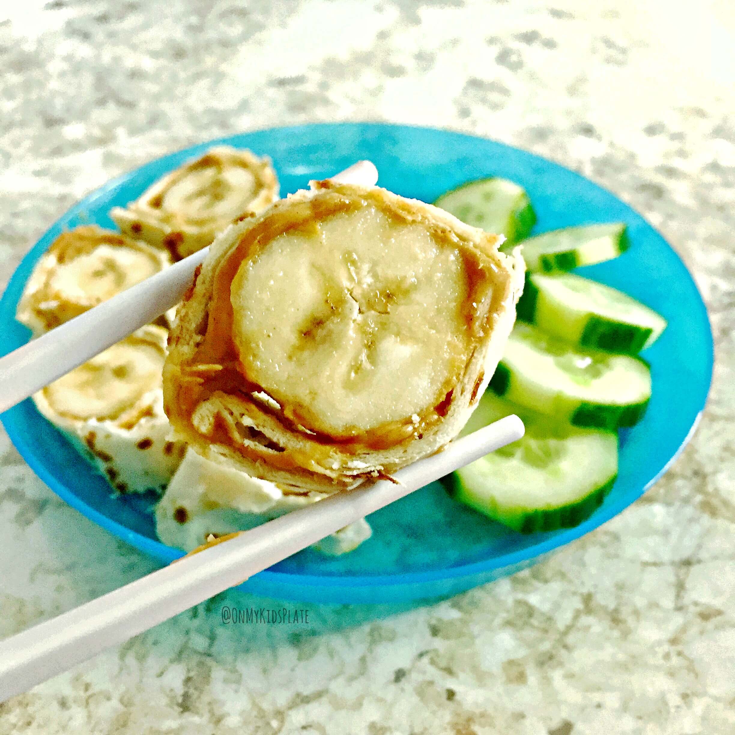 We see a close up of a blue kid's plate as it sits on a kitchen counter full of banana wrapped in a tortilla with a peanubutter inside wrapped and sliced into round pieces of babana sushi. A pair of chopsticks holds one slice of banana sushi up for a close look. A side of sliced cucumbers are stacked as a side ont he plate for a quick snack. 