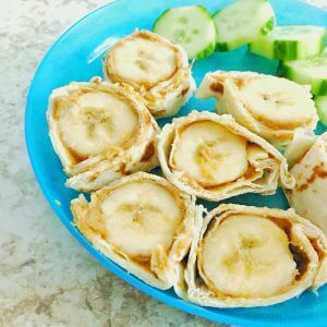 banana sliced with peanut butter wrapped in tortilla on a plate with sliced cucumber