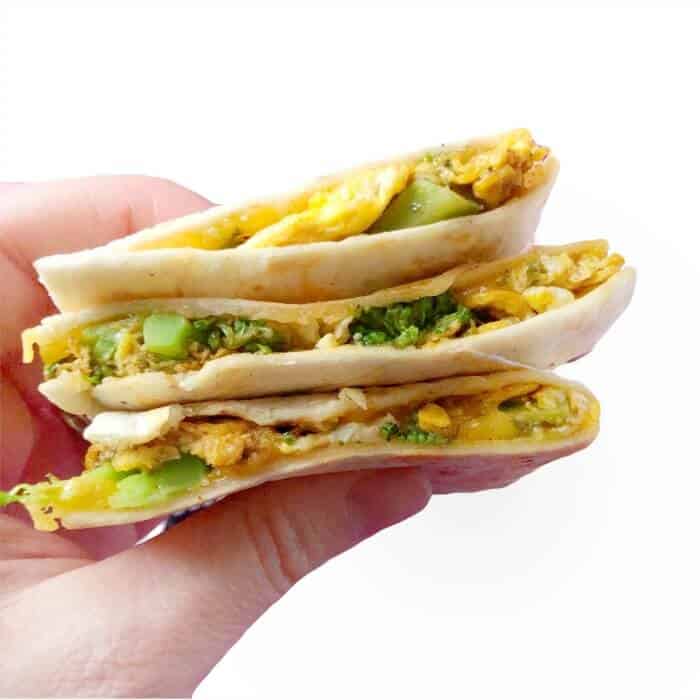 Three pieces of quesadilla stacked being held by a hand stuffed with egg, cheese and broccoli 
