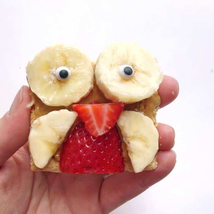 A hand holding a snack of banana slices and strawberries on a cookie shaped like an owl