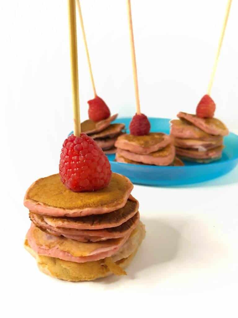 Mini pink raspberry pancakes on kabobs, one in the front, and three in the background on a plate. Each mini pancake stackis topped with a raspberry.