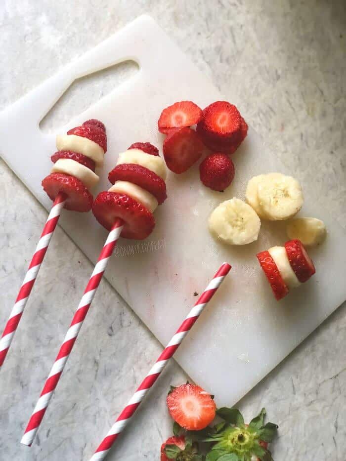 Strawberry and banana slices on a cutting board being added to sticks to look like the cat in the hat\'s hat.