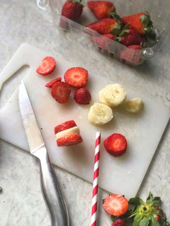 Strawberries and bananas being sliced with a knife on a cutting board