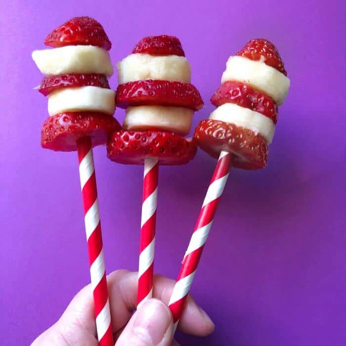 Three red and white sticks are held up with strawberry and banana slices stacked on the end alternating to look like Dr. Seuss Cat in the Hat's hat as a birthday treat.