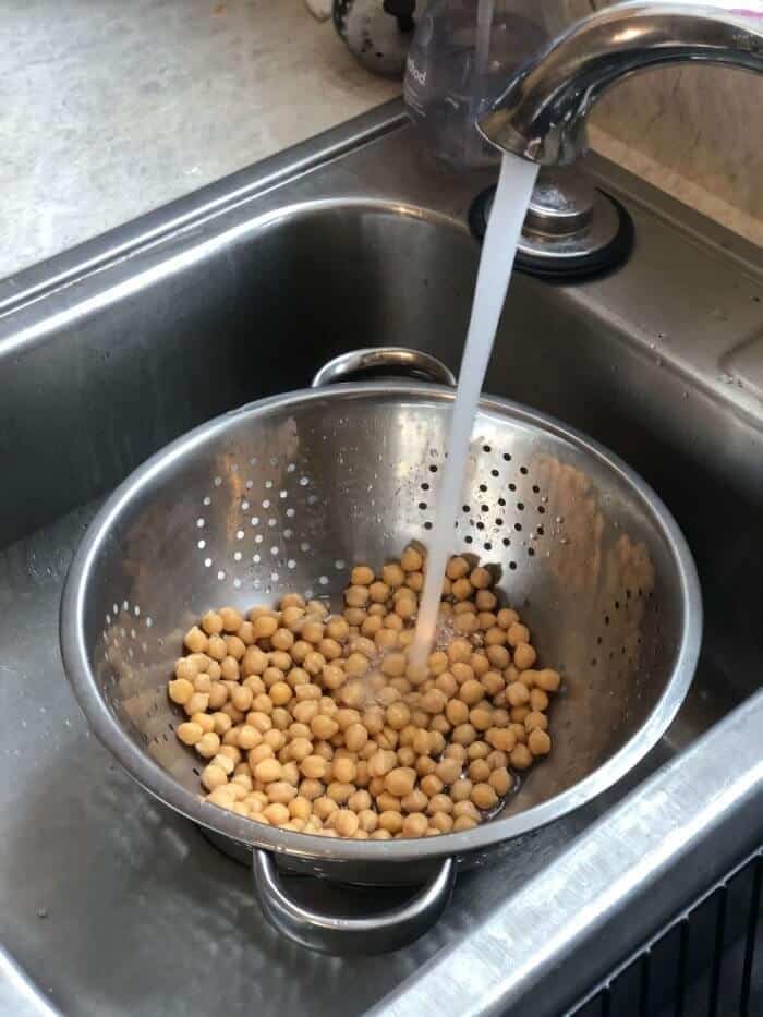 A collander sits in a sink full of chickpeas being rinsed.