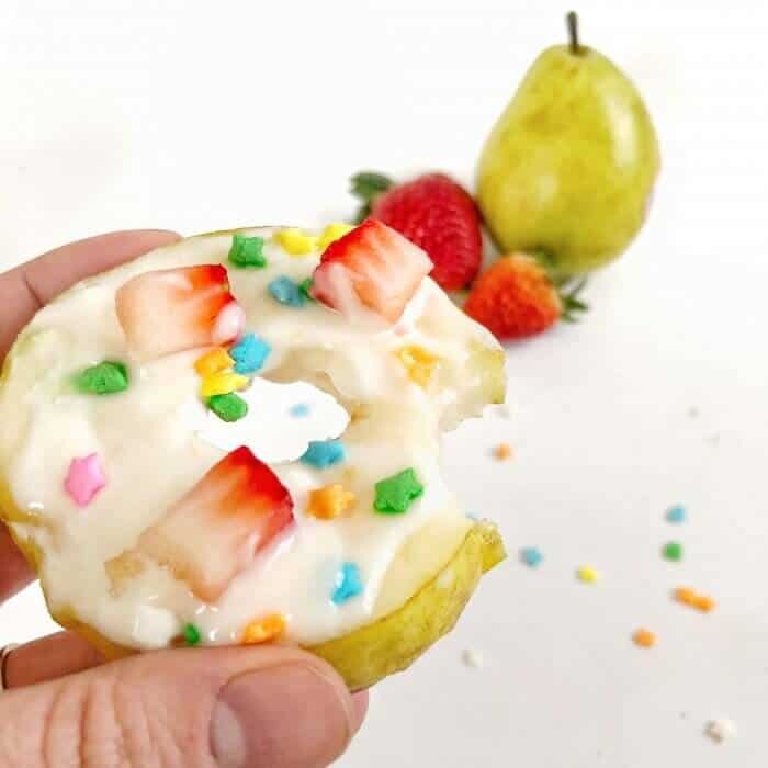 A pear decorated with sprinkles and fruit to look like a donut with fruit behind
