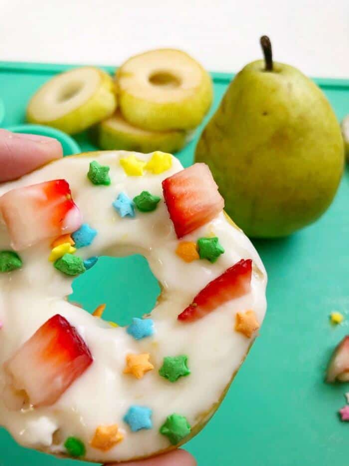 Close up of a pear slice decorated with yogurt, sprinkles and fruit pieces to look like a donut with more pear in the background