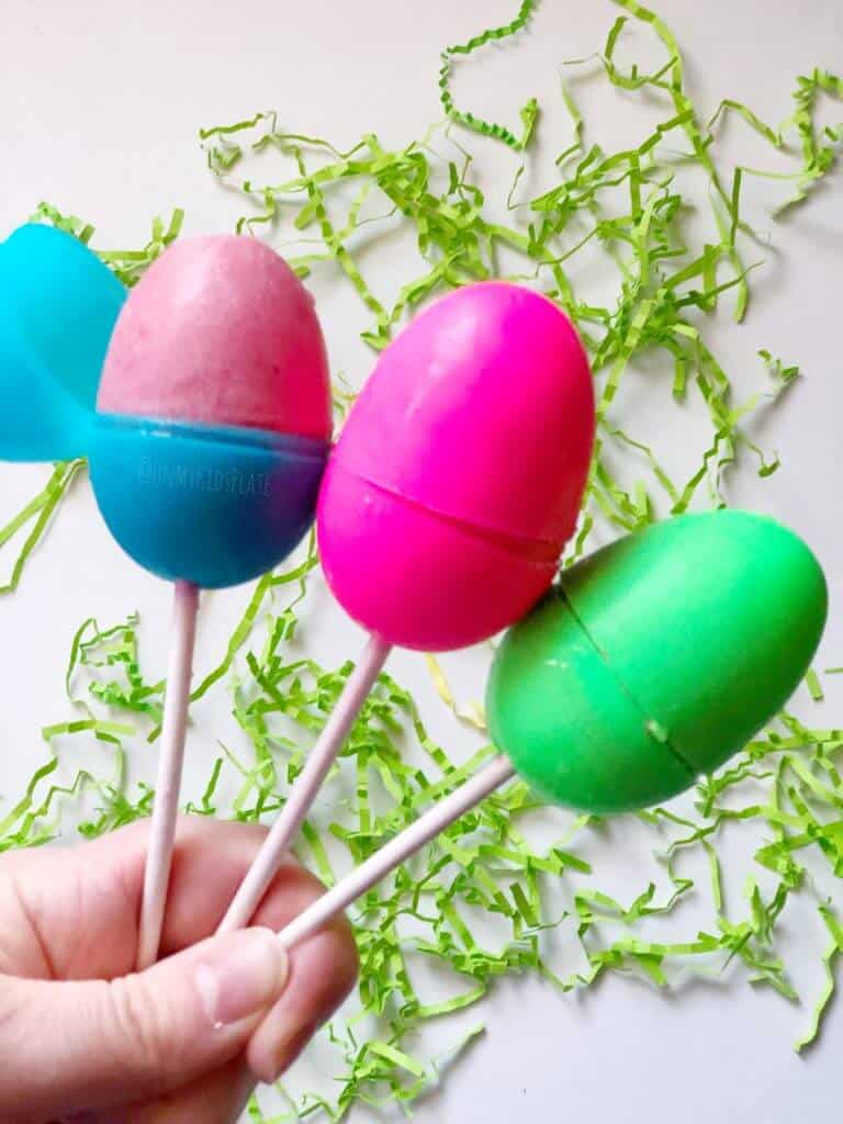 Three Easter eggs on top of sticks filled with frozen popsicle filling are held by a hand in front of a table with scattered Easter grass behind.