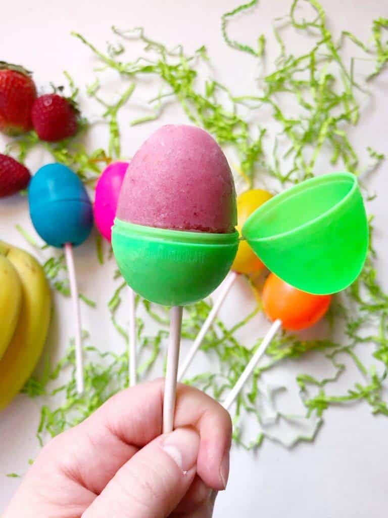 A hand holds an open Easter Egg on a stick full of frozen strawberry raspberry popscile. Behind the Easter Egg popsicles is several more laying on a table among scattered green Easter grass. Strawberries and banananas can be seen on the left side of the image.