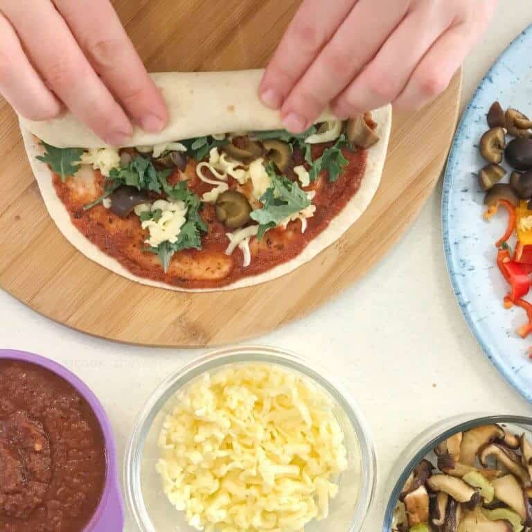 A tortilla full of red sauce, cheese and olived being rolled with ingredients in bowls nearby