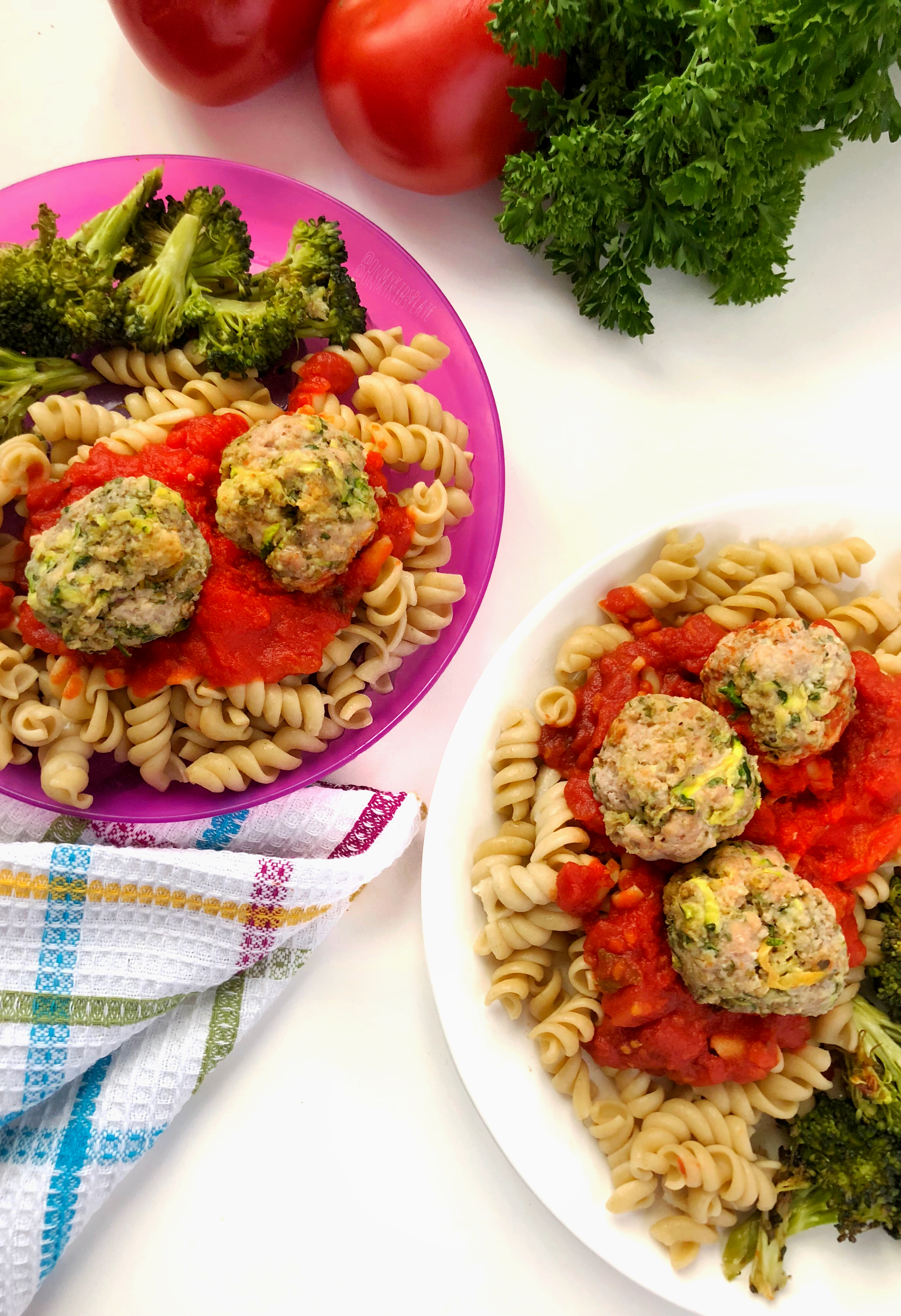 Two plates of pasta with meatballs and broccoli, one is a white plate and one is a purple child\'s plate