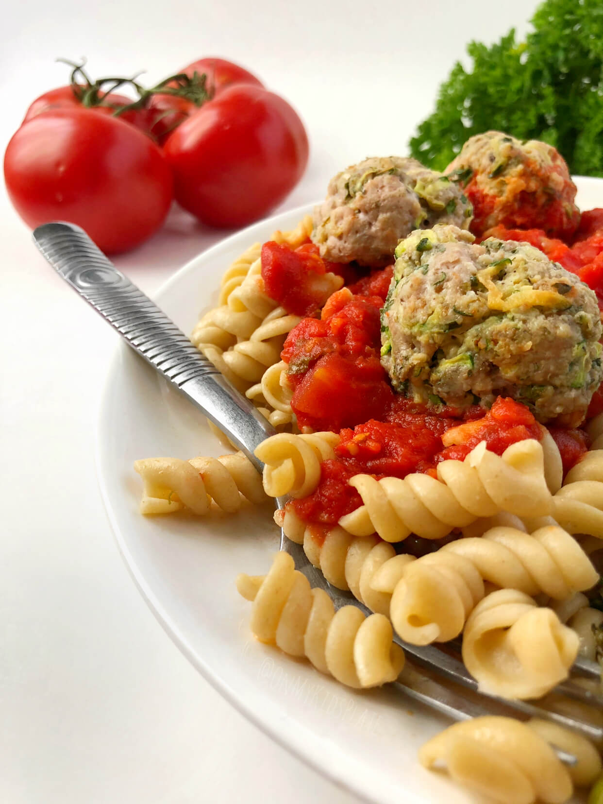 A plate of meatballs, pasta and tomato sauce with tomatoes and parsely in the background