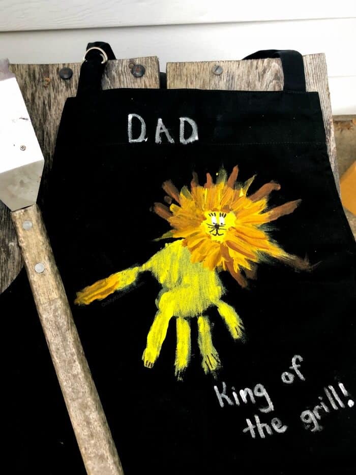 A lion painted on an apron close up made from a child\'s handprint wit a grill brush laying next to it.