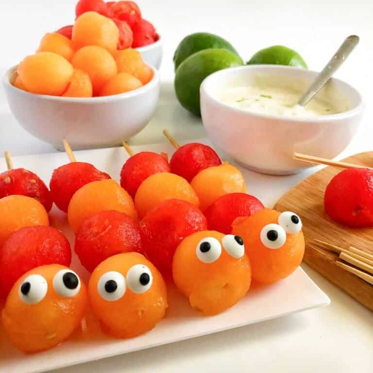 Melon balls decorated to look like caterpillars on sticks with more fruit and yogurt behind