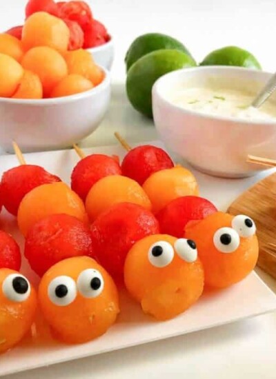 Melon balls decorated to look like caterpillars on sticks with more fruit and yogurt behind