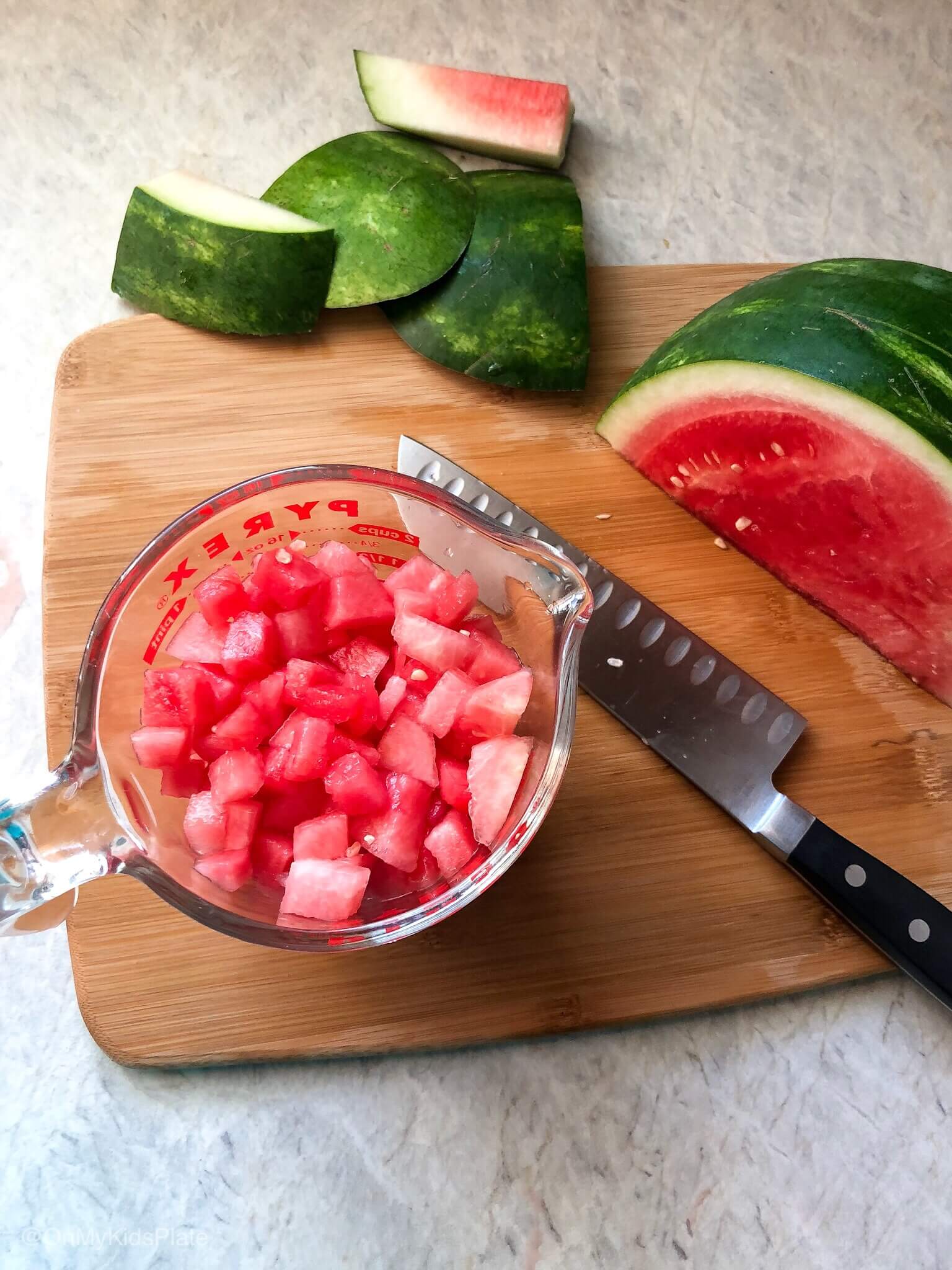 Watermelon diced in a measuring cup next to a knife and a piece of watermelon on a cutting board.