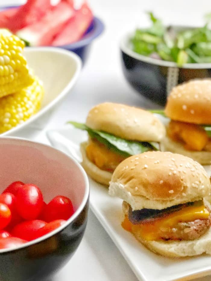 A platter of turkey burgers sits next to a summer picnic meal of tomatoes, corn and salad.