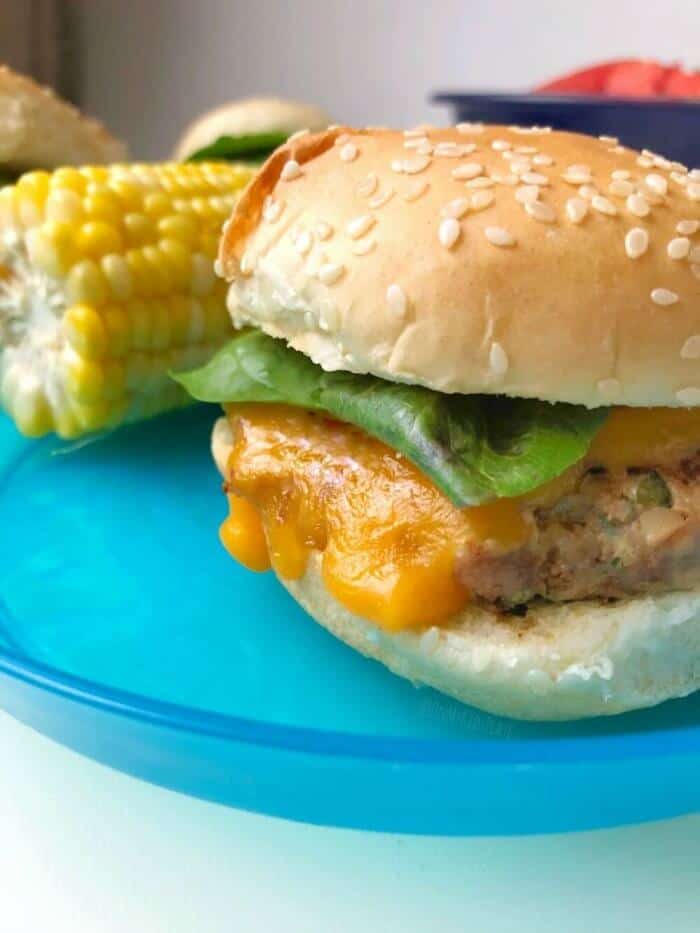 A turkey slider with cheese sitting on a kid\'s plate next to a piece of cooked corn on the cob.