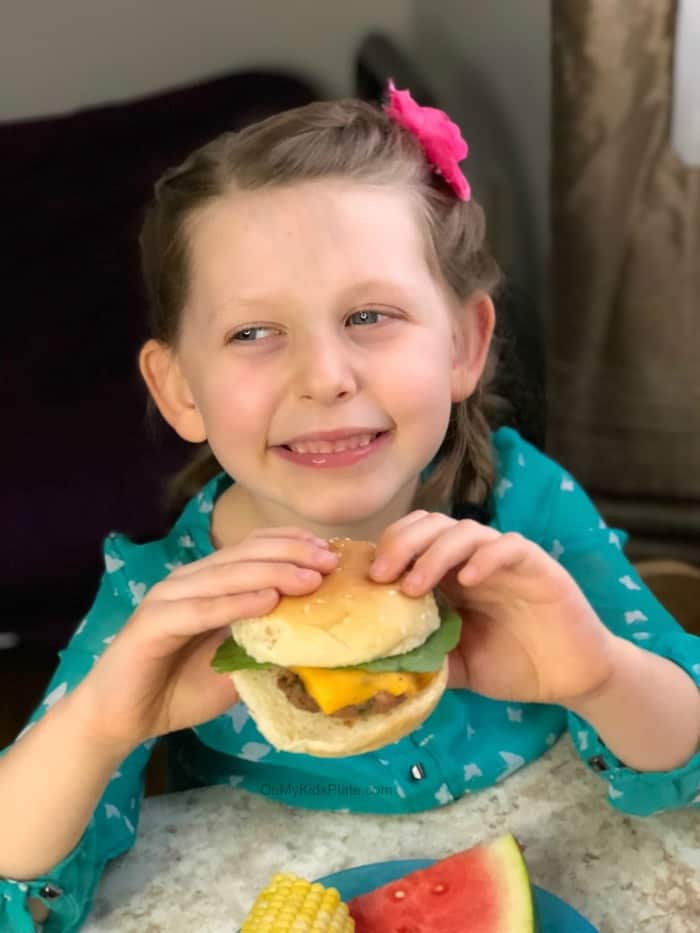 Little girl holds a turkey burger slider with cheese with both hands while smiling.