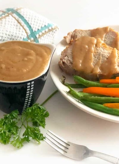 A container of brown gravy in a small gravy boat with a plate of pork, gravy and vegetables next to it