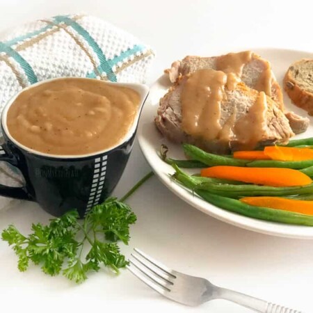 A container of brown gravy in a small gravy boat with a plate of pork, gravy and vegetables next to it