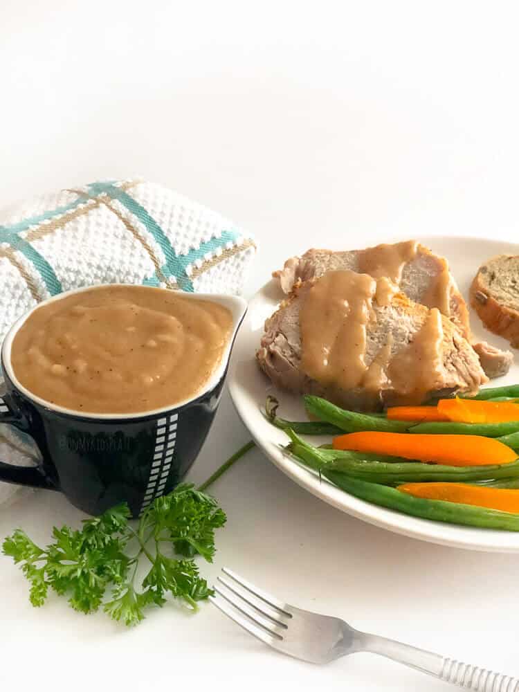 A container of brown gravy in a small gravy boat with a plate of pork drizzled in gravy, and green beans and peppers next to it.