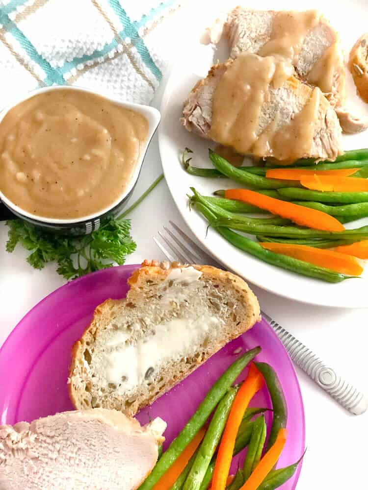 A child and an adult plate of pork, vegetables and bread next to a container of brown gravy