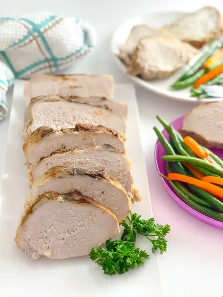 Delicious boneless pork loin recipe from the crock pot is sliced on a platter, surrounded by plates full of pork with vegtables for a tasty dinner.
