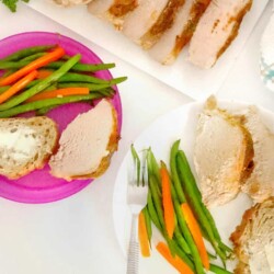 A platter of pork loin sliced next to an adult and kids purple dinner plate with vegetables and bread
