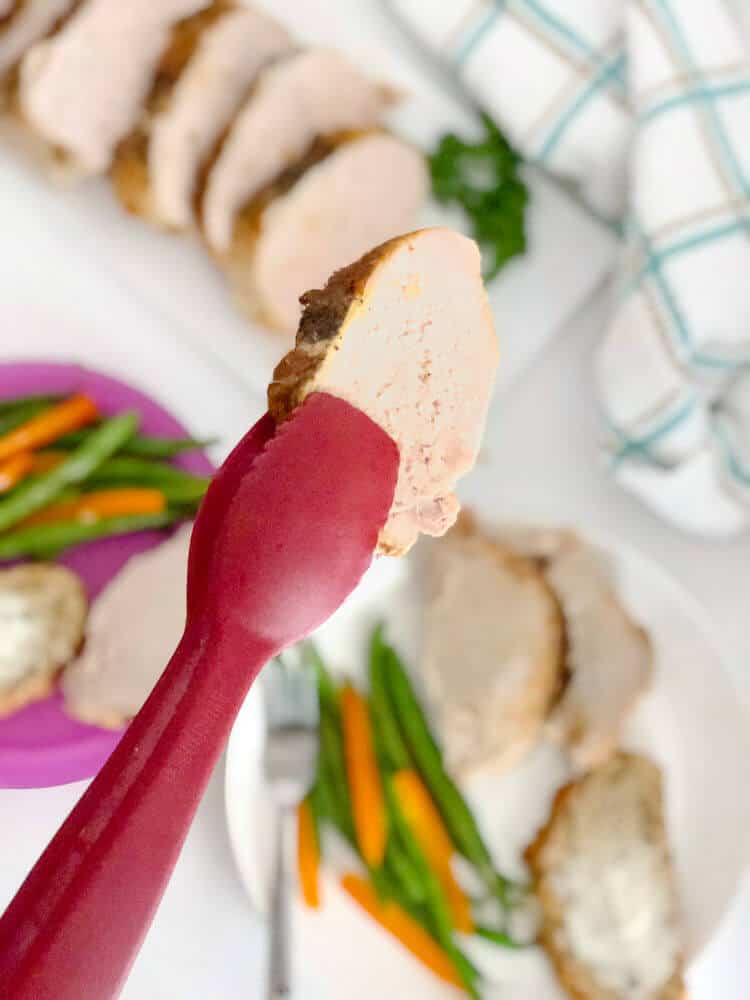A slice of boneless pork loin is held up by tongs over a family meal with vegetables plated.