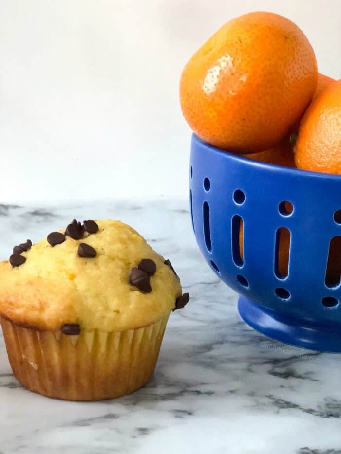 A bowl of oranges on a table next to an orange muffin