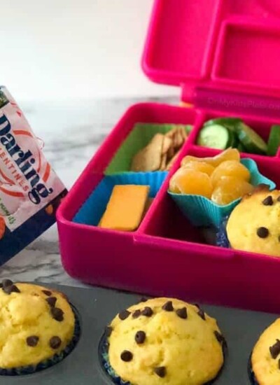 A child's lunchbox full of food with muffins in a pan in  front and a bag of clementines in the background