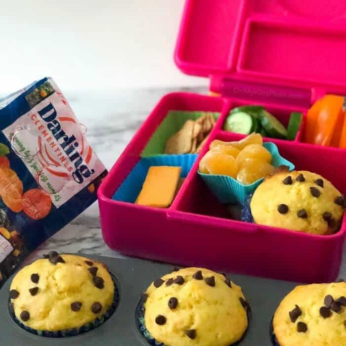 A child's lunchbox full of food with muffins in a pan in  front and a bag of clementines in the background