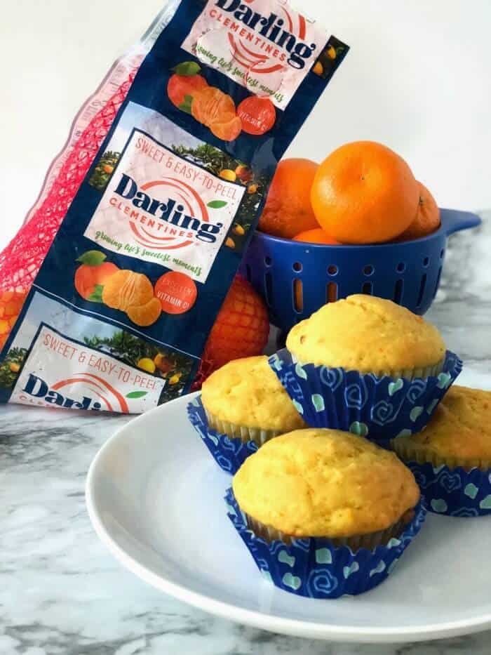 A plate of orange muffins with a bowl and container of clementines from the grocery store behind