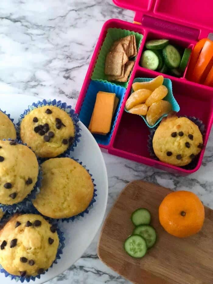 A child\'s lunchbox from overhead next to a stack of muffins and a cutting board where food is being prepped.