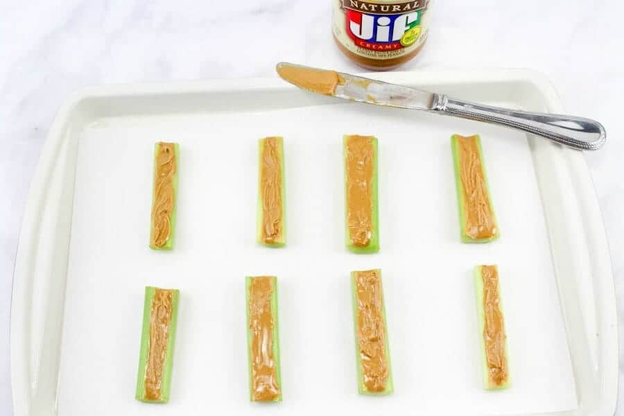 Eight slices of celery are on a platter covered in peanut butter. Above the celery is a jar of peanut butter and a knife covered in nut butter.