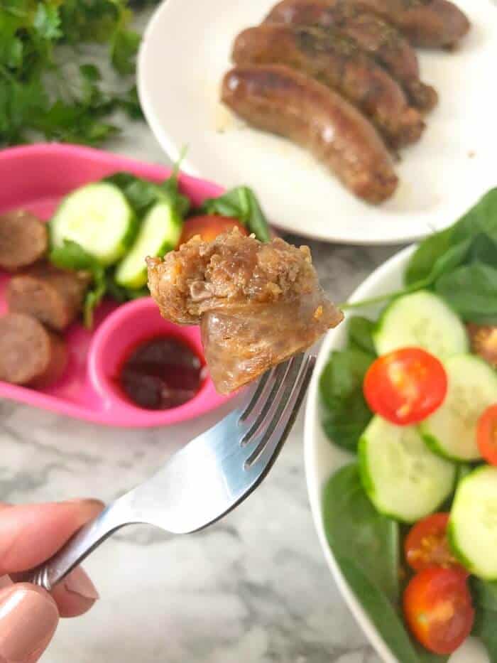A piece of sausage being held on a fork over a plate of sausage and a plate of salad