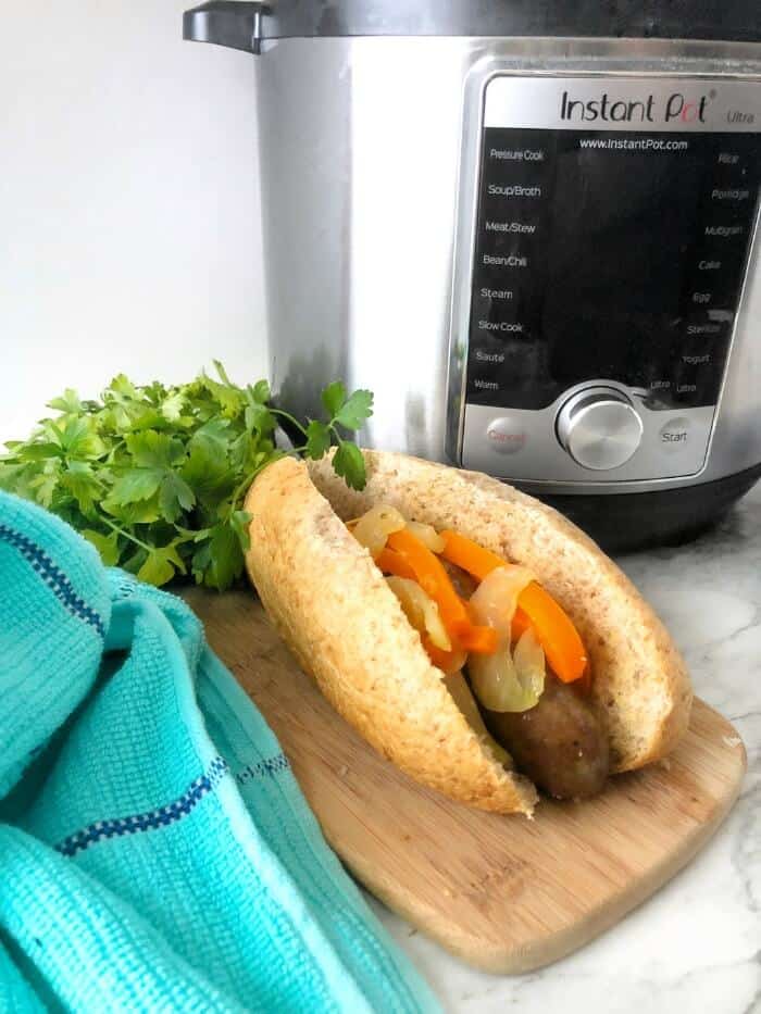 A sausage in a bun topped with peppers and onions on a cutting board next to an instant pot