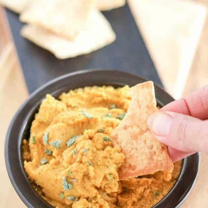 pumpkin hummus in a bowl being scooped with pita bread by a hand.