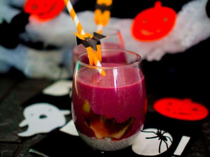 A dark purple smoothie decorated with a bat straw and more Halloween decorations on the table nearby.