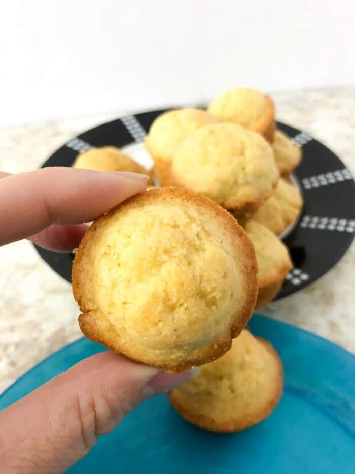 A hand holds a cornbread muffins close up to the camera over a little kid's plate, in front of a serving plate full of muffins.