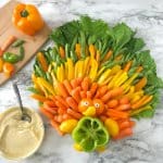 On the table is a vegetable platter shaped like a turkey with orange, yellow and green feathers made out of bell peppers, carrots, sugar snap peas and lettuce. In the center of the platter is a bell pepper cut to look like a turkey but arranged that you can fill the pepper with dip. A side of hummus with a spoon also rests on the table and a cutting board with several cut bell pepper pieces sits nearby.