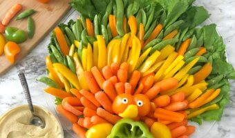 On the table is a vegetable platter shaped like a turkey with orange, yellow and green feathers made out of bell peppers, carrots, sugar snap peas and lettuce. In the center of the platter is a bell pepper cut to look like a turkey but arranged that you can fill the pepper with dip. A side of hummus with a spoon also rests on the table and a cutting board with several cut bell pepper pieces sits nearby.