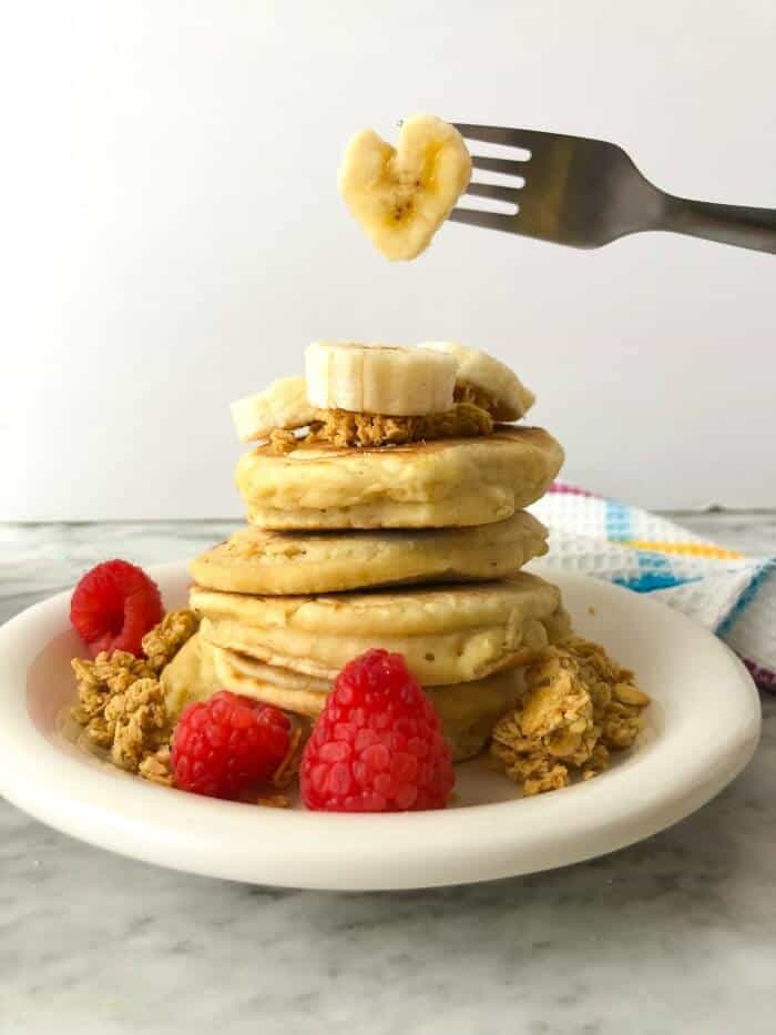 Large stack of fluffy pancakes with bananas and raspberries. A fork holds a heart shaped piece of banana above.