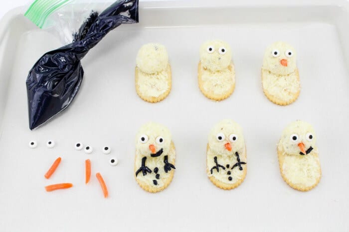 Cheese crackers being decorated with carrots, candy eyes and black cream cheese to make snowmen