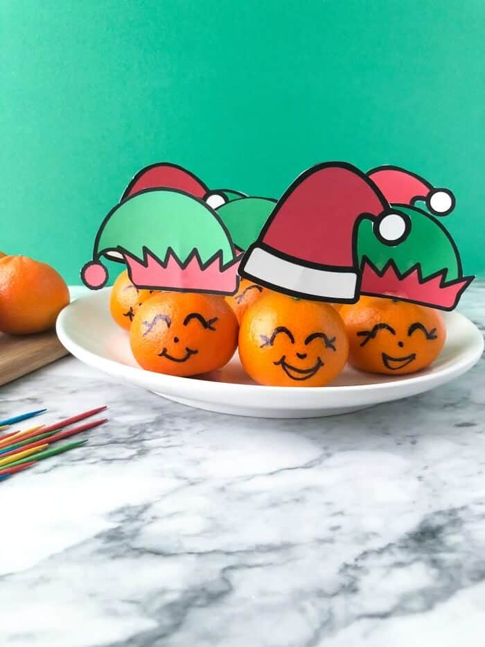  A bunch of clementines on a plate all wearing santa and elf hats with faces drawn on. Toothpicks and another clementine sit next to them