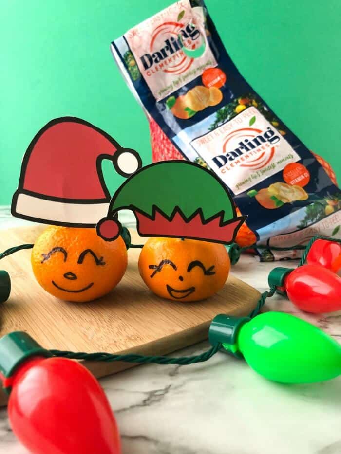Clementines with faces drawn on and wearing paper santa and elf hats next to a bag of clementines